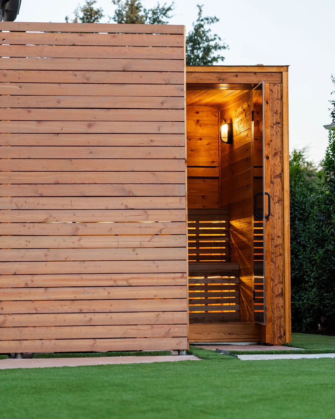 Open glass door revealing glowing backlit slat benches inside cedar sauna