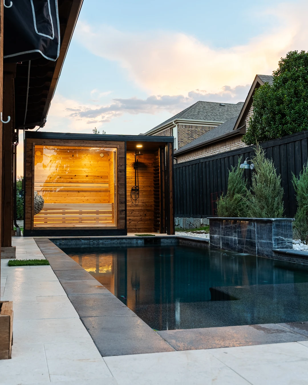 Charred-dark sauna cube glowing beside a backyard pool at dusk