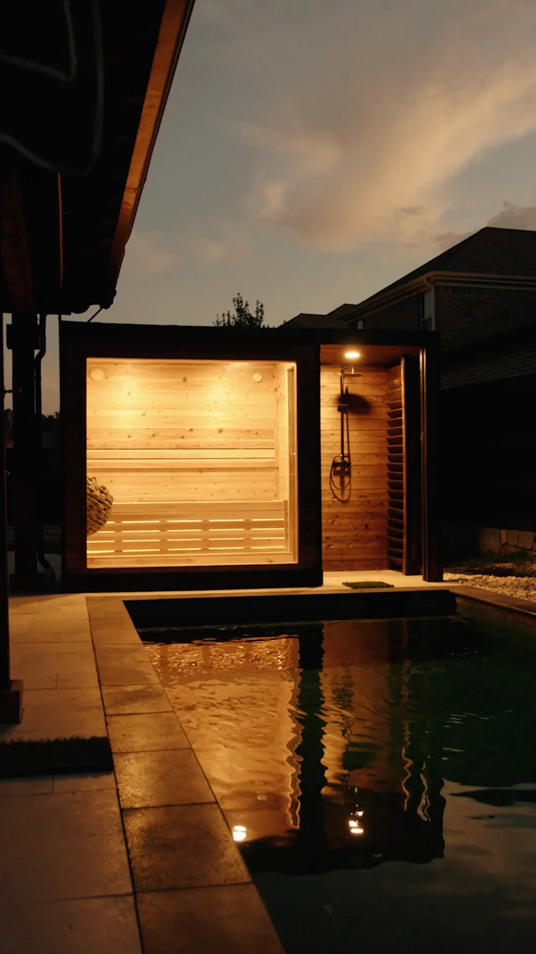 Charred-dark sauna cube glowing beside a backyard pool at dusk